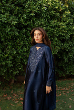 Load image into Gallery viewer, Woman wearing a dark blue dress with intricate patterns standing in front of green foliage.
