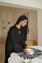 تصویر گیلری ویور میں لوڈ کریں، A woman with long black hair wearing a black embroidered kurta and matching dupatta sits at a white table, holding a striped bowl on a black-and-white patterned runner in a bright, modern room.
