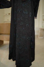 تصویر گیلری ویور میں لوڈ کریں، A close-up of a black long-sleeved dress with dark blue floral embroidery and tassel details, shown on a model in a softly lit indoor setting.
