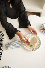 تصویر گیلری ویور میں لوڈ کریں، A person wearing a black embroidered dress sits at a white table, holding a ceramic teacup on a striped saucer. A matching striped napkin or small cloth lies nearby on the table.
