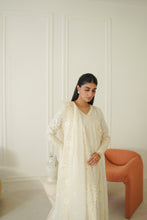 Load image into Gallery viewer, A woman wearing a cream-colored embroidered traditional outfit with a matching dupatta, standing in a bright room with a white paneled wall and an orange chair.
