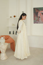 Load image into Gallery viewer, A woman wearing a long, flowing cream-colored embroidered dress stands in a bright, modern white room with minimalist decor, including an orange chair and a side table.
