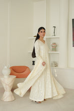 Load image into Gallery viewer, A woman wearing a long, flowing white embroidered dress with tassel details, standing in a bright, modern white room with decorative shelves and a small orange chair.
