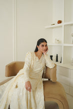 Load image into Gallery viewer, A woman wearing a cream-colored embroidered traditional outfit sits on a beige armchair in a bright, modern room with white walls and built-in shelves.
