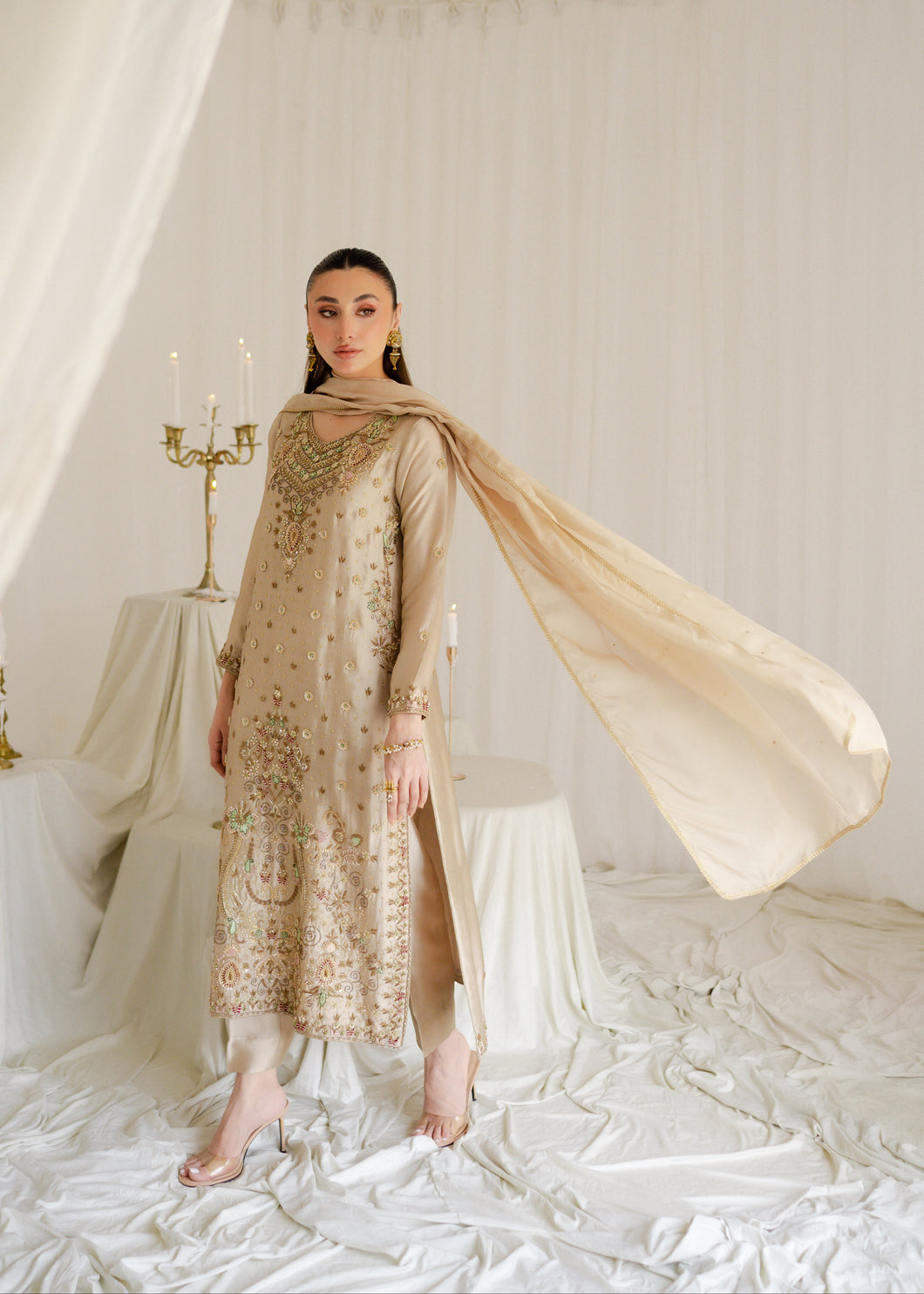Woman in a gold embroidered outfit with a beige scarf in a white, draped setting.